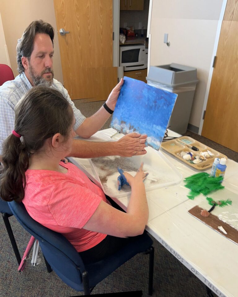 A volunteer holds up a finished blue and white ocean canvas with a textured sandy shoreline, while an artist presses their hand into a sand-filled tray on the table.