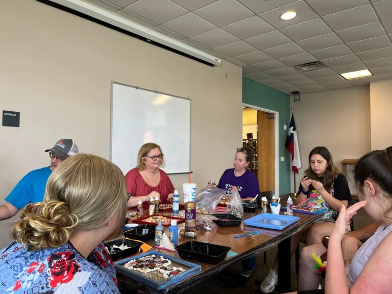 A wide view of the cork art table, with artists and volunteers gluing corks and beads onto framed boards, craft supplies spread across the tabletop.