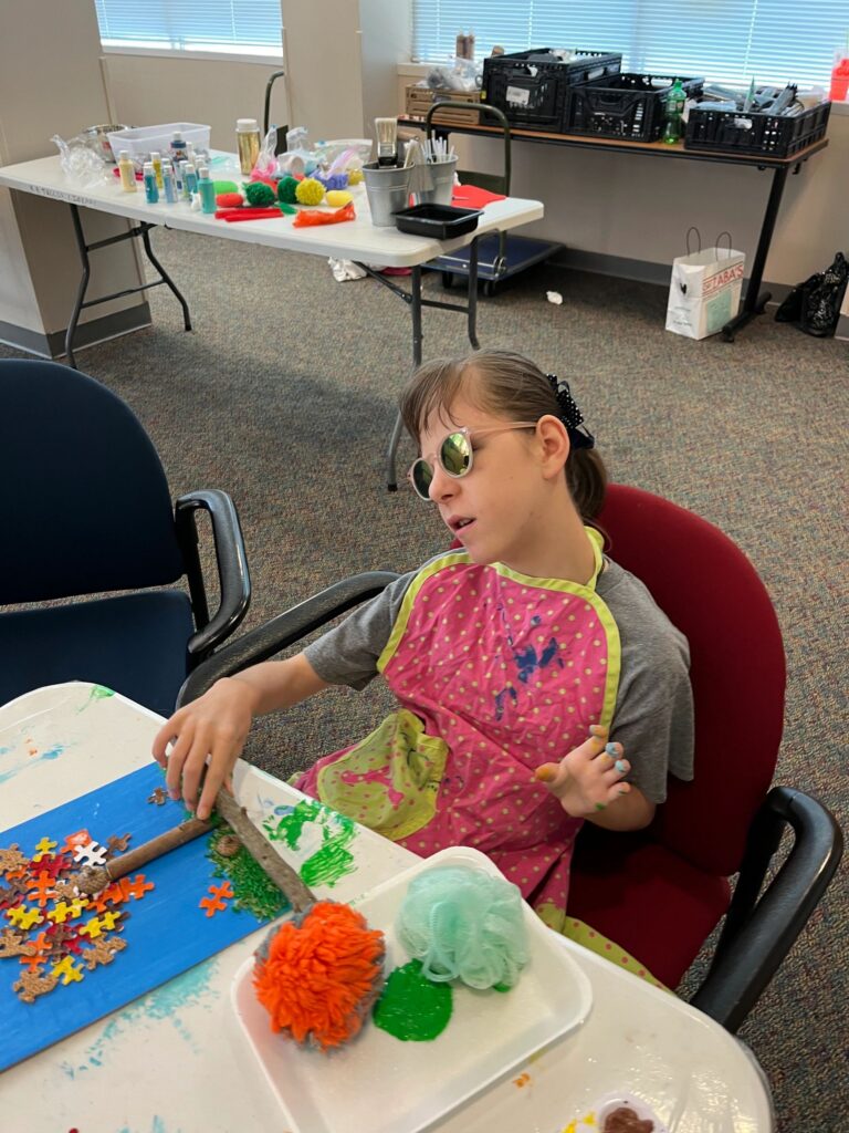 An artist scoops green paint from a tray onto a canvas with a twig trunk, tray of colourful pom-poms and a green scrubber nearby on the table.