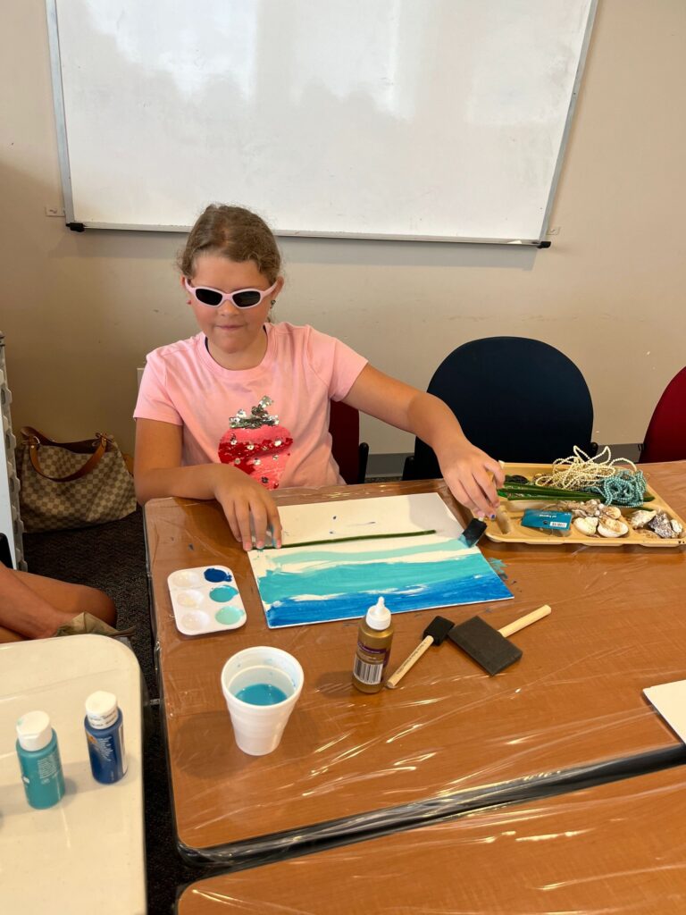 An artist paints a two-tone blue ocean scene on a canvas, with a tray of beaded strands, shells and paint supplies on the table.