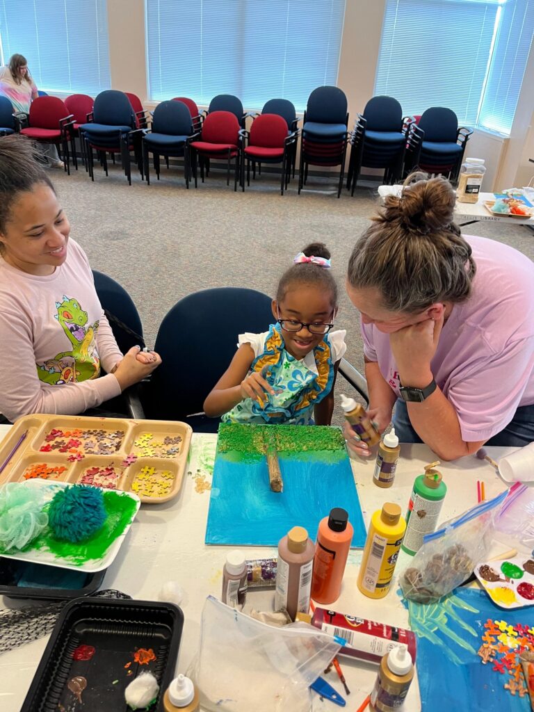A volunteer helps an artist glue puzzle pieces onto a canvas already painted with a green and blue landscape and a twig trunk, with a paint tray of yellow and gold puzzle pieces nearby.