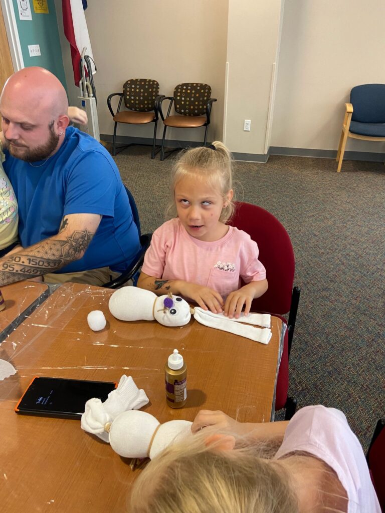 An artist holds a white sock bunny decorated with a blue glitter butterfly and a pink pom-pom nose, seated at a table.