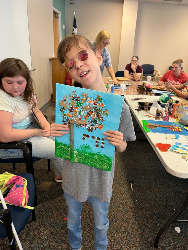 An artist holds up a fall tree canvas with a canopy of puzzle pieces in red, orange and gold over a twig trunk, with green sprinkled grass at the base.