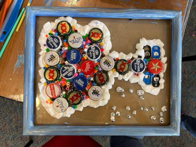 A framed cork piece shaped like a bird, with pull-tabs forming the head and beads scattered at the base against a foam backing.