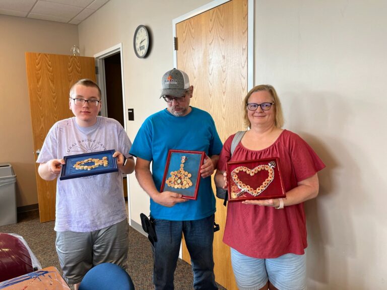 Three artists hold up their finished frames side by side: one with cork letters spelling DAD, one with a cork airplane design, and one with a heart made from painted corks.