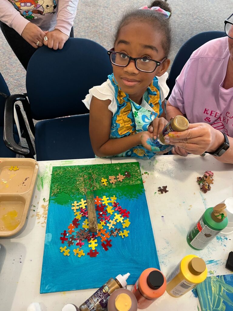 An artist holds up a canvas showing a tree made from gingerbread-coloured puzzle pieces over a twig trunk, with green sprinkled grass and a yellow sun.