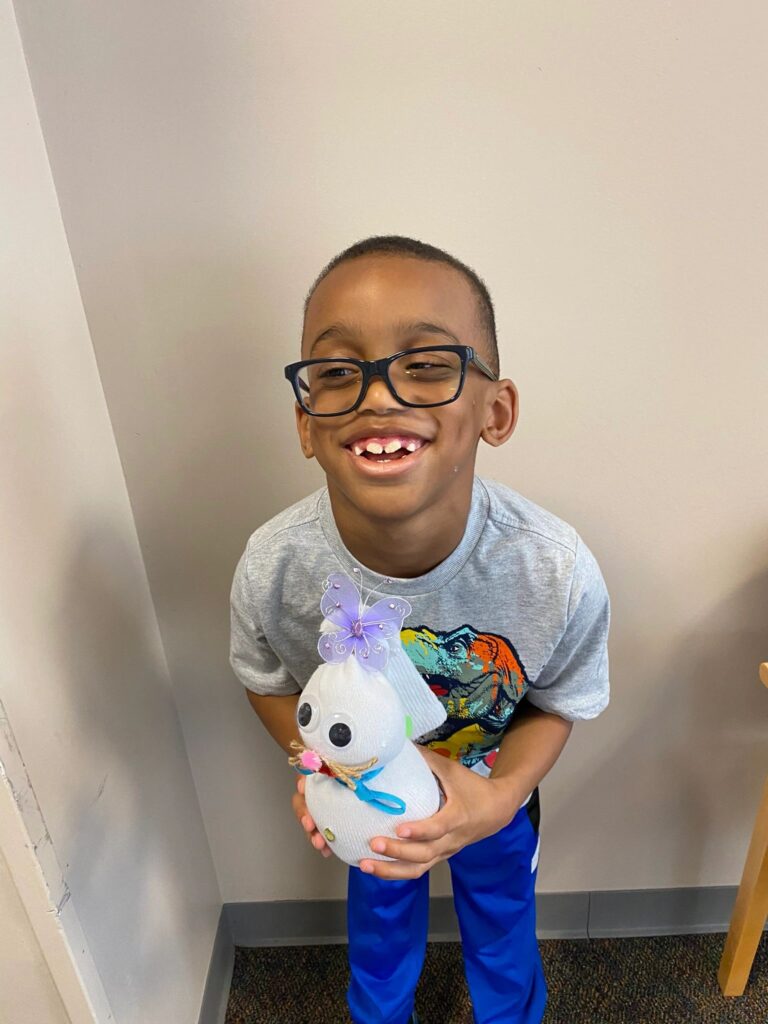 A young artist smiles while holding a finished sock bunny with a purple butterfly, googly eyes and a pink pom-pom nose.