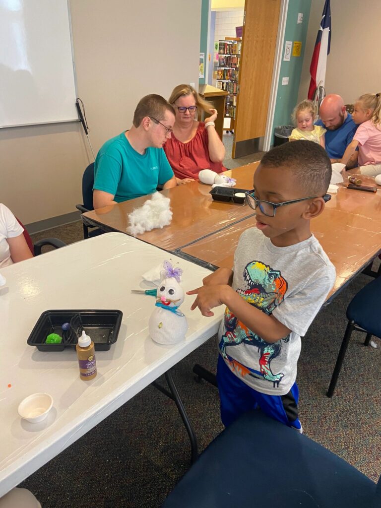 A volunteer talks with a young artist at a table set with a finished sock bunny, glue and a bowl of rice, other artists working nearby.