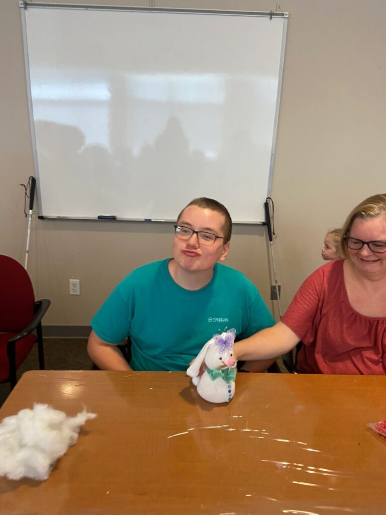 An artist sits at a table with a finished sock bunny decorated with a purple butterfly and green ribbon bow, a volunteer smiling beside them.