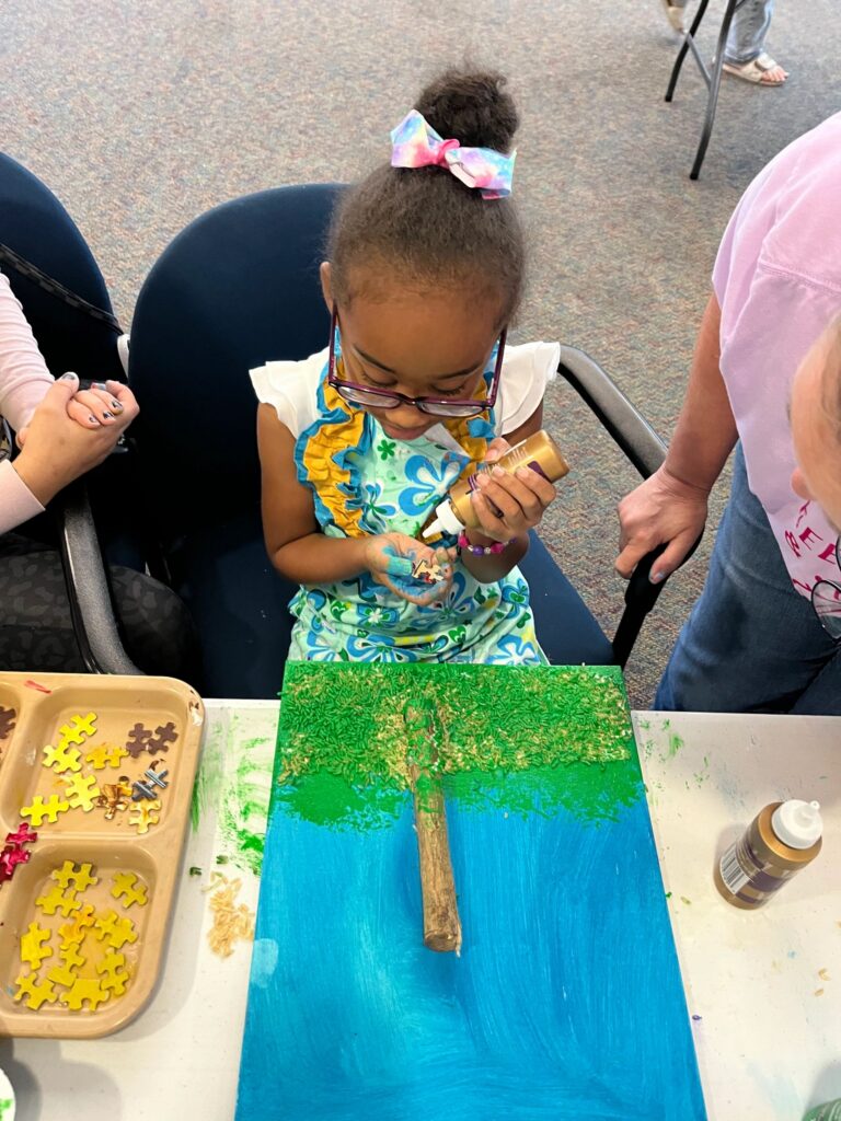 A volunteer helps an artist paint blue and gold onto a canvas already decorated with a twig trunk and green sprinkled grass.