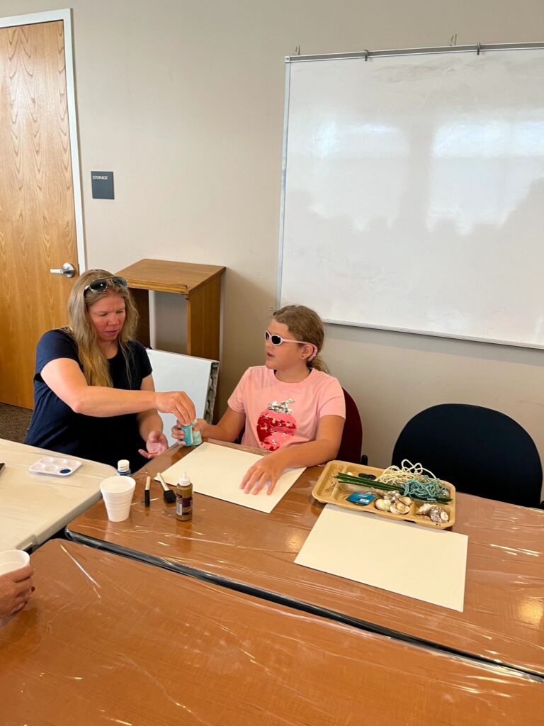 A volunteer helps an artist glue small shells onto a cream-coloured canvas, with a tray of shells, netting and beaded strands on the table.