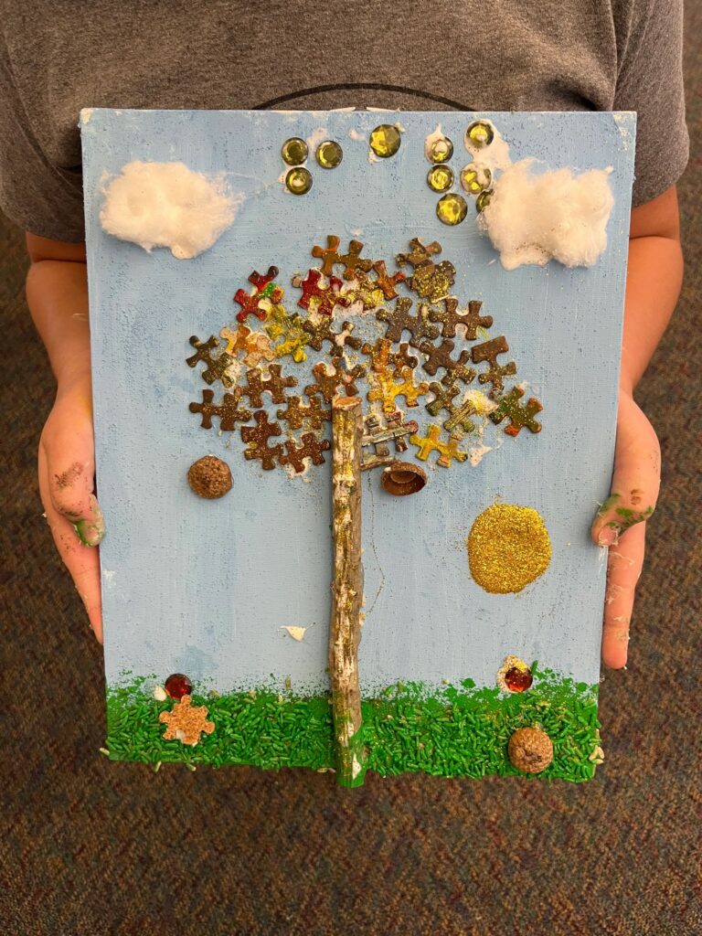 A fall tree canvas held up close: puzzle pieces in bronze, gold and red form the leafy canopy around a real twig trunk, with cotton-wool clouds, green sprinkled grass and gold glitter suns in the sky.