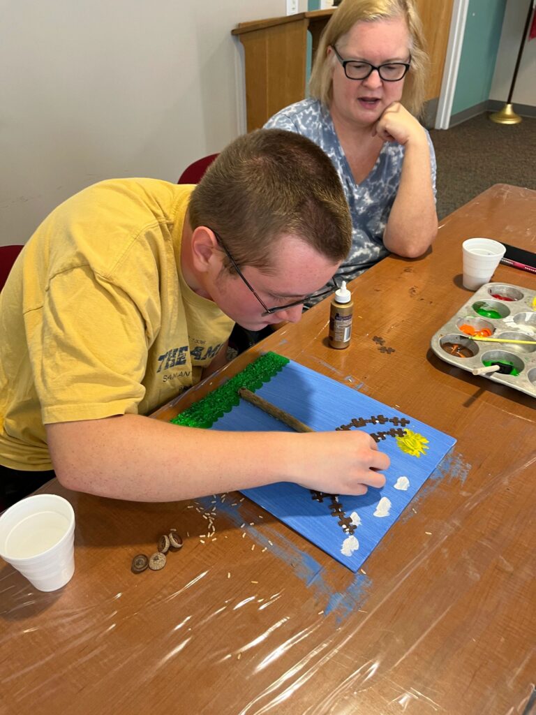 A volunteer looks on as an artist presses puzzle pieces and acorns onto a blue and green canvas with a twig trunk.