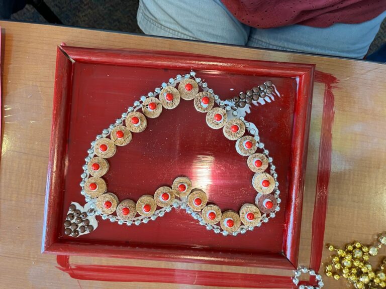A framed cork piece in the shape of a heart, made from cork slices with red bead centres and a rhinestone border, resting on a table.