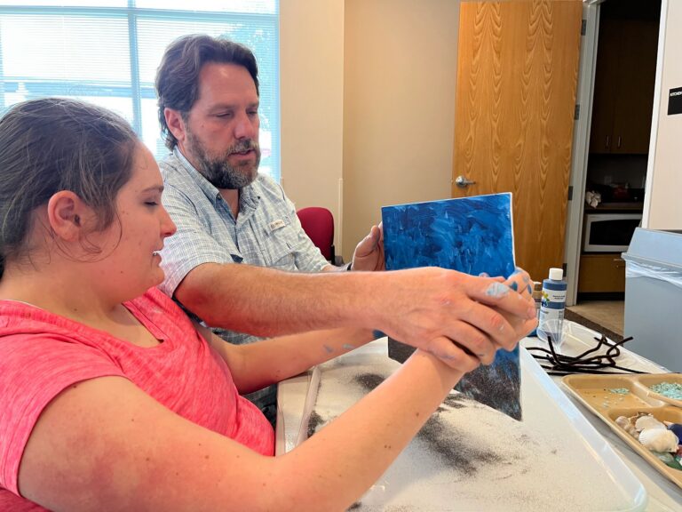 A volunteer helps an artist paint a blue canvas with sand texture, with beaded strands and shells laid out on a tray on the table.