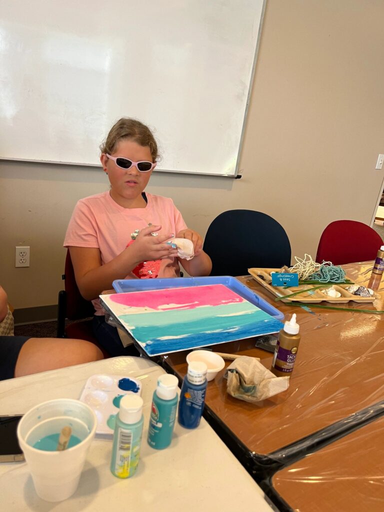 An artist glues shells onto a canvas already dotted with a cluster of small rhinestones, with a tacky glue bottle and scissors on the table.