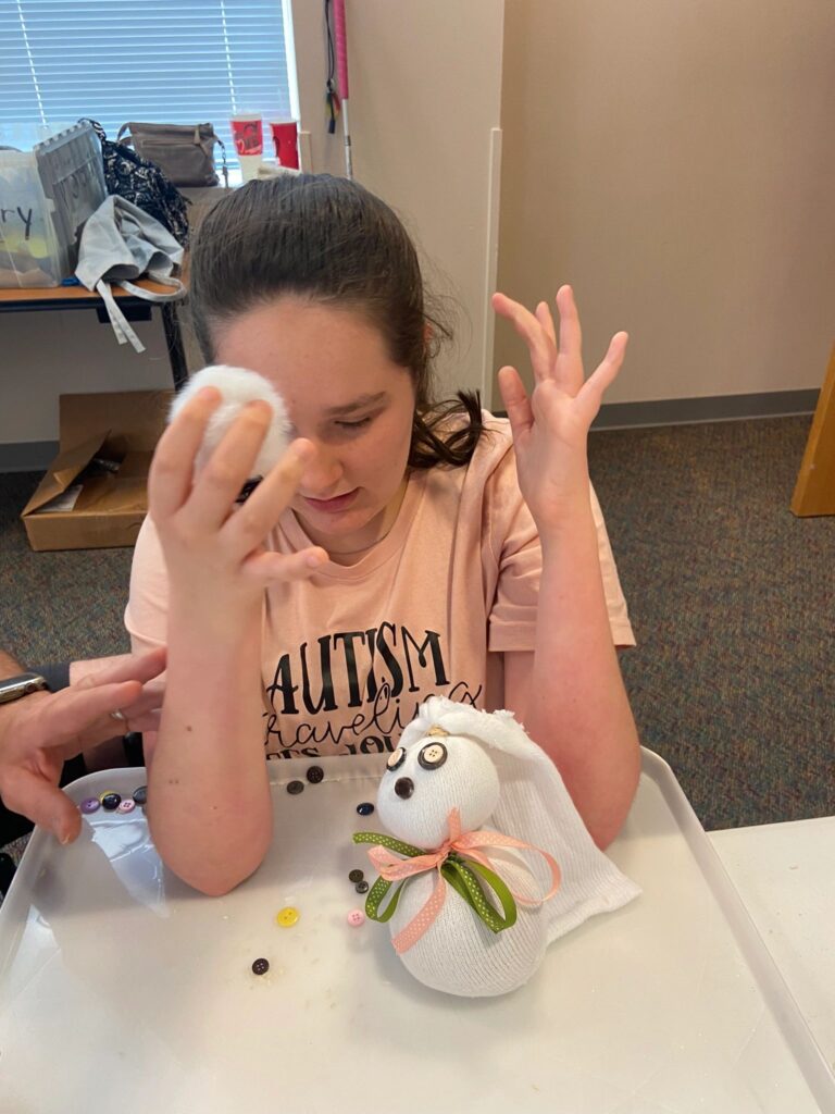 An artist holds a cotton-wool ball near a finished sock bunny decorated with a button face and ribbon bow, a volunteer helping alongside them.