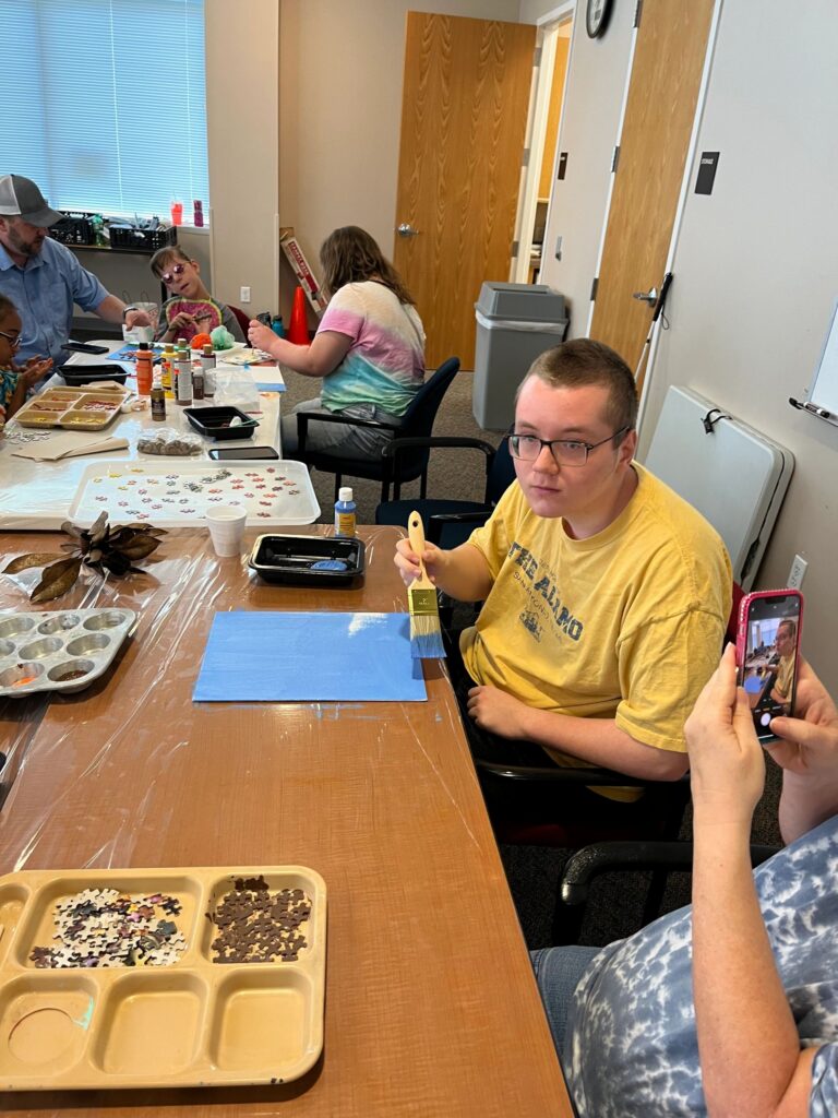 A volunteer and an artist look at a canvas together, the artist's hand resting on a scattering of gingerbread-coloured puzzle pieces near a twig trunk.