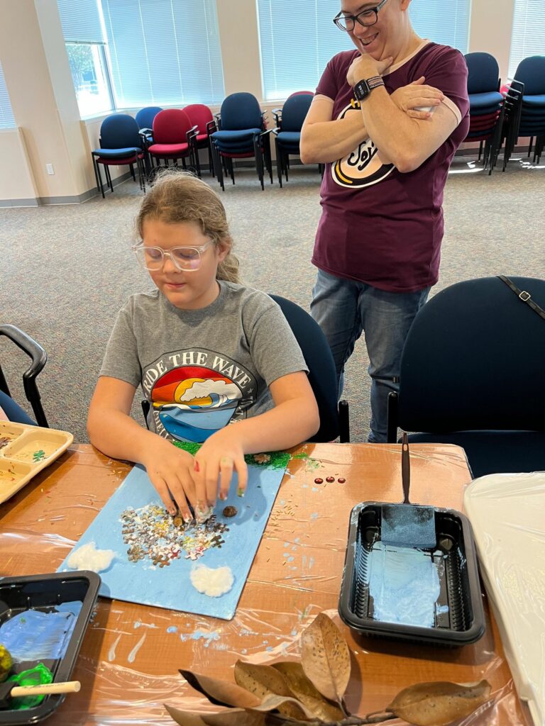 An artist presses a mix of puzzle pieces and small gems into wet glue on a blue canvas at a table set with paint trays and dried magnolia leaves.