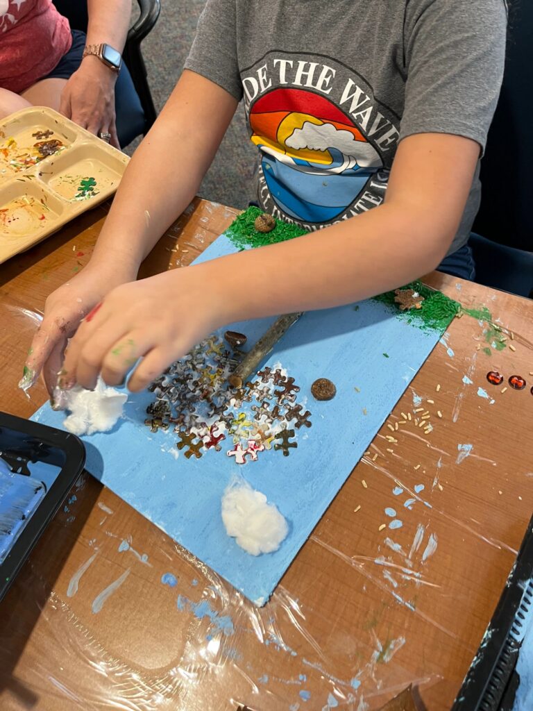 An artist scoops white glue onto a blue canvas already dotted with dark puzzle pieces, acorns and small red gems, with green grass strips at each side and a twig trunk across the middle.