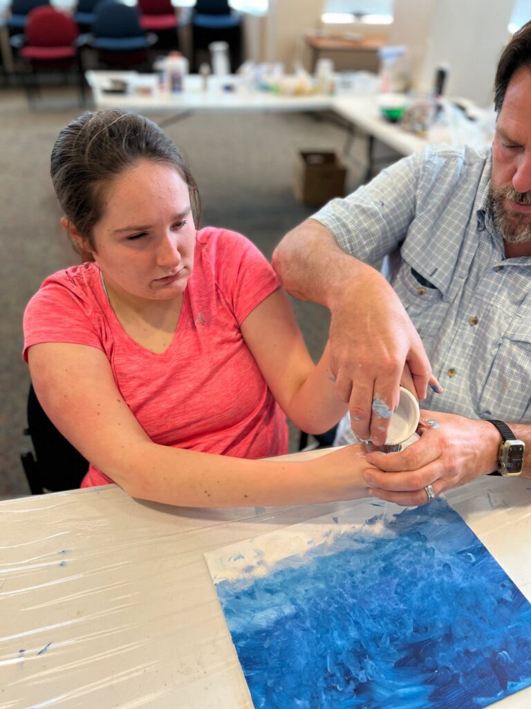 A volunteer helps an artist blend white paint powder onto their hands, with a finished blue canvas on the table.