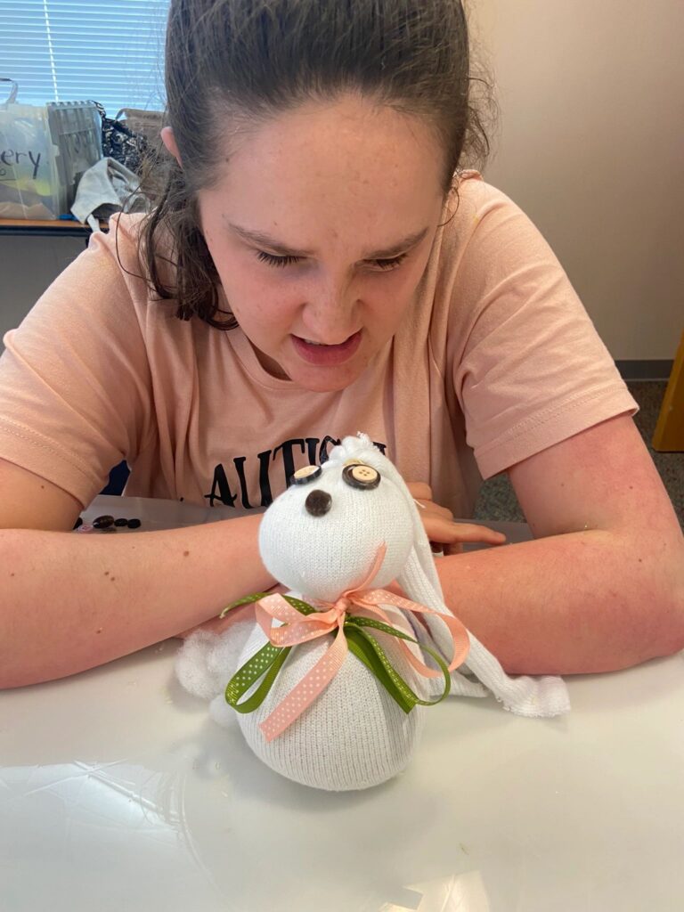 An artist looks closely at a finished sock bunny decorated with buttons and a ribbon bow, resting on the table in front of them.