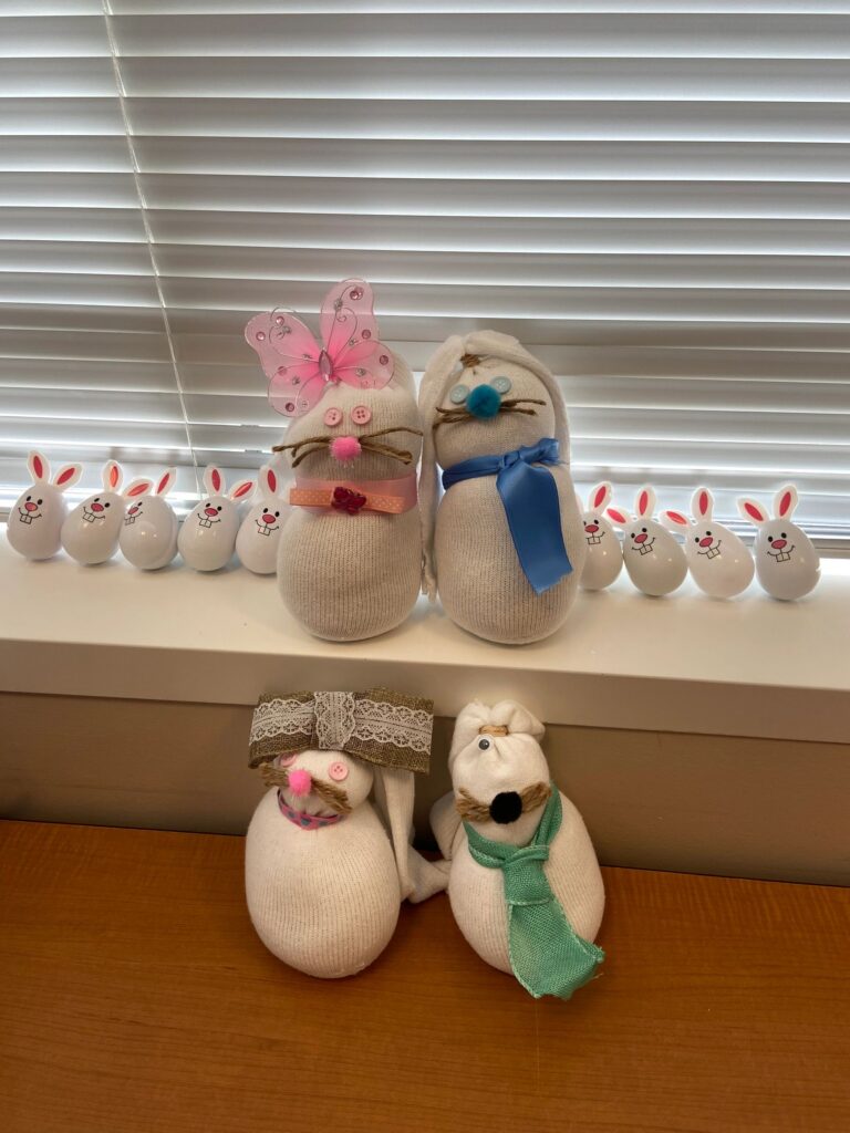 A row of finished sock bunnies lined up on a windowsill, decorated with butterfly wings, ribbon bows and pom-pom noses, alongside a set of small painted bunny figurines.