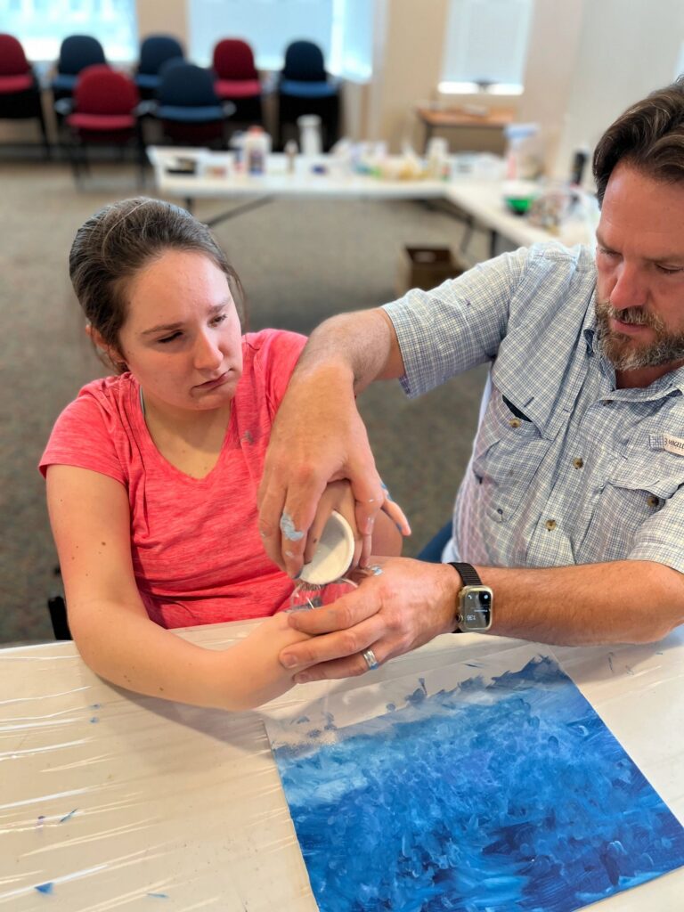 A volunteer helps an artist blend white paint powder onto their hands over a finished blue canvas.