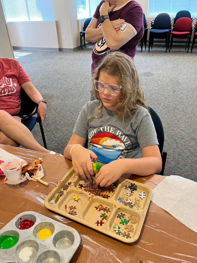 An artist sorts pink, red and green puzzle pieces at a divided tray, working at a table set with paint trays.