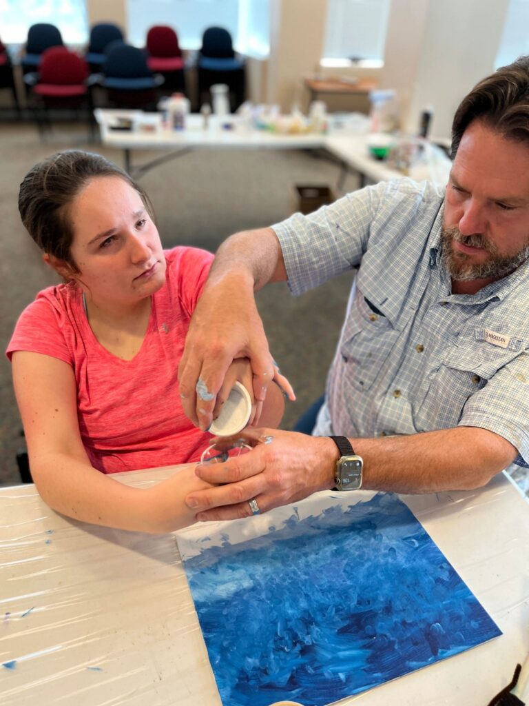 A volunteer helps an artist spread white paint powder over their hands, with a finished blue canvas resting on the table between them.