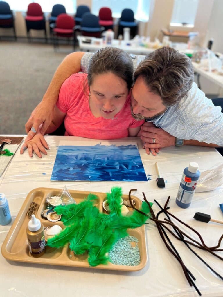 A volunteer and an artist lean close together over a finished dark blue ocean canvas, with a tray of green feathers, shells and beaded strands nearby.