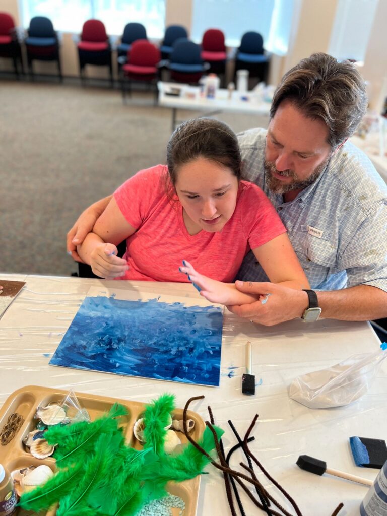 A volunteer wraps an arm around an artist as they both look closely at a finished blue and white canvas, with a tray of shells and green feathers on the table.
