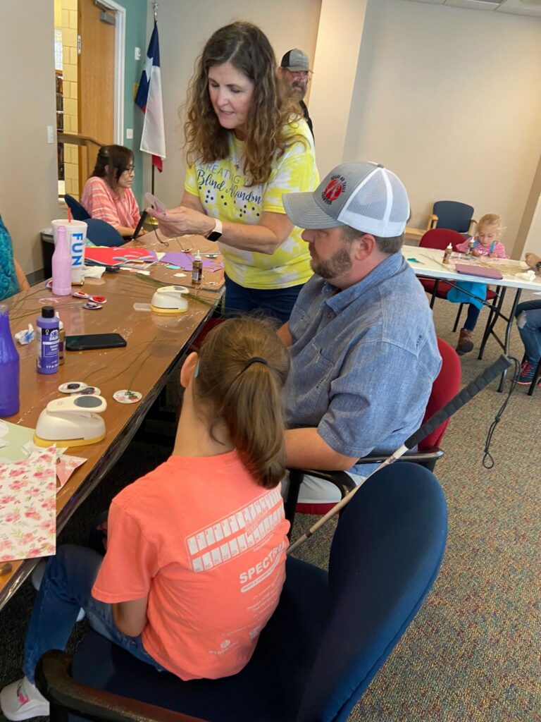 A volunteer shows a small purple charm to an artist, with tables of sock bunnies, glue and craft supplies around them.