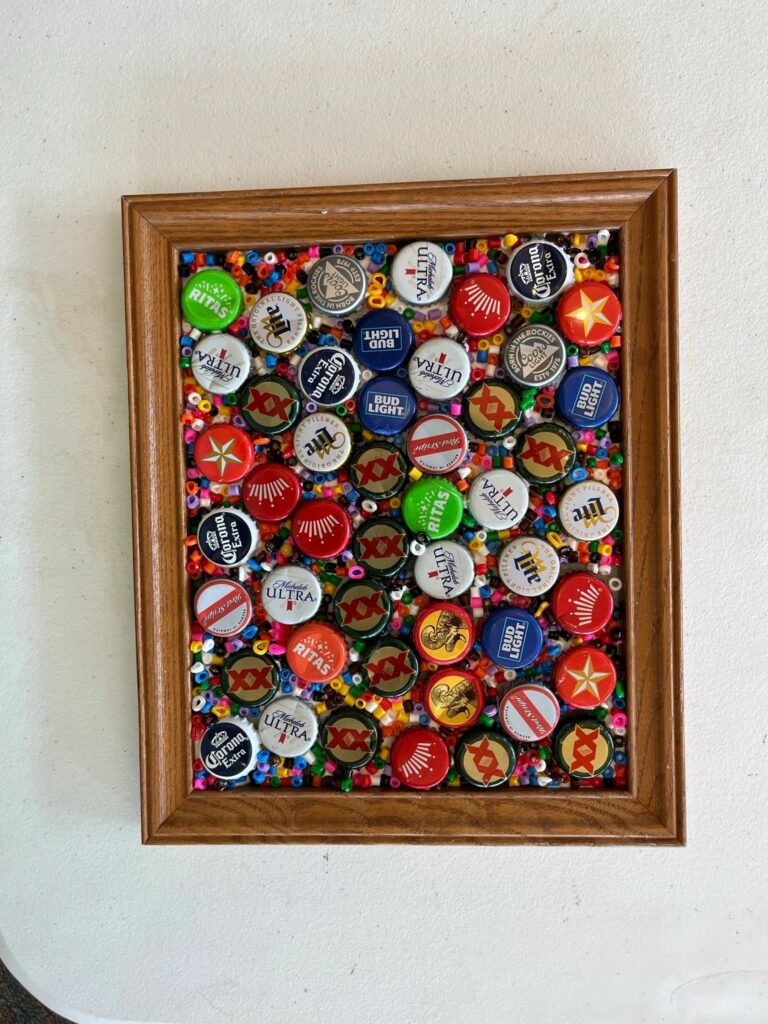 A framed piece covered edge to edge with bottle caps in red, white, blue and green, nestled among colourful pony beads.