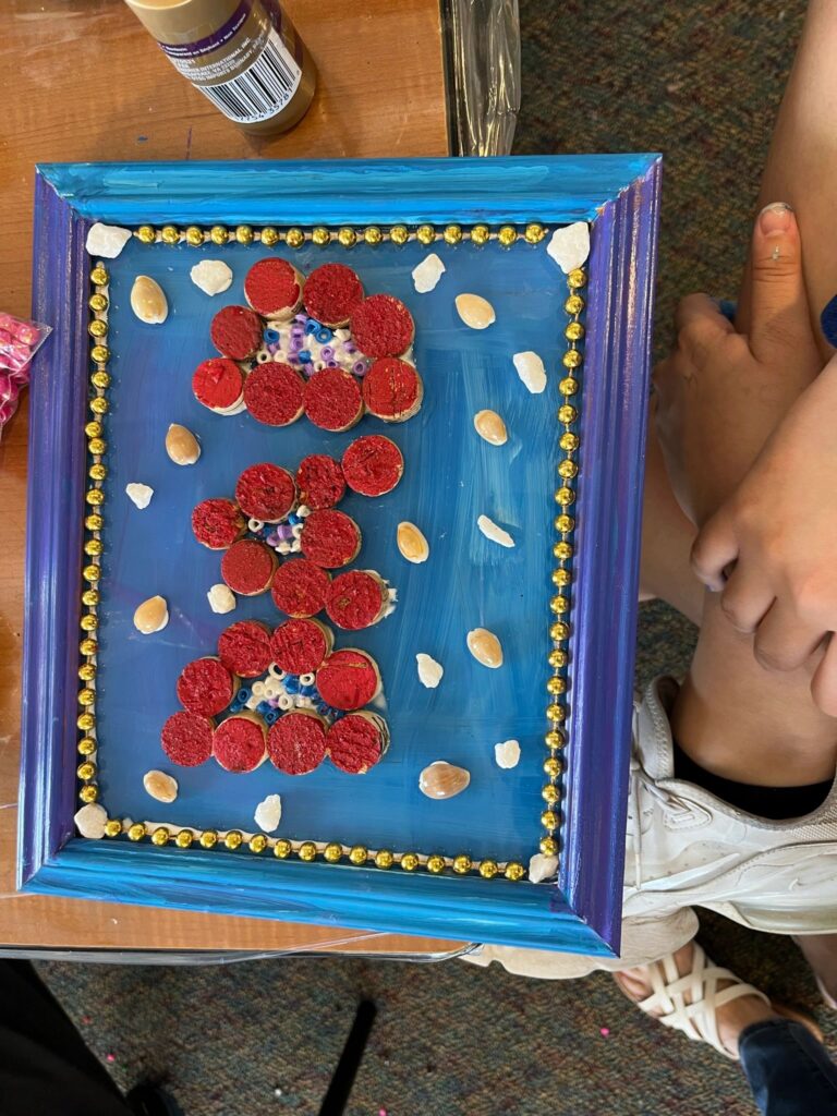 A framed piece with three flower shapes made from red-painted cork slices in a row, bordered by gold beads and small shells on a blue background.
