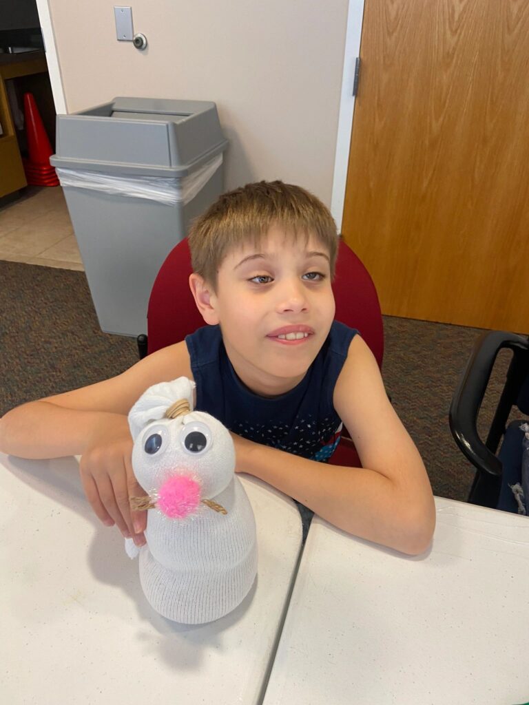 An artist holds a finished sock bunny with big googly eyes, a pink pom-pom nose and an orange button, smiling for the camera.