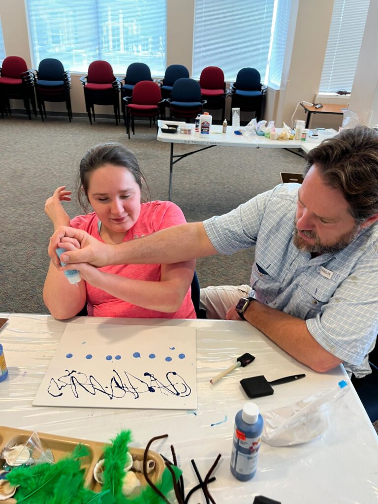 A volunteer helps an artist trace lines of blue paint dots on a canvas, forming letters that spell a word, with green feathers and shells nearby on the table.