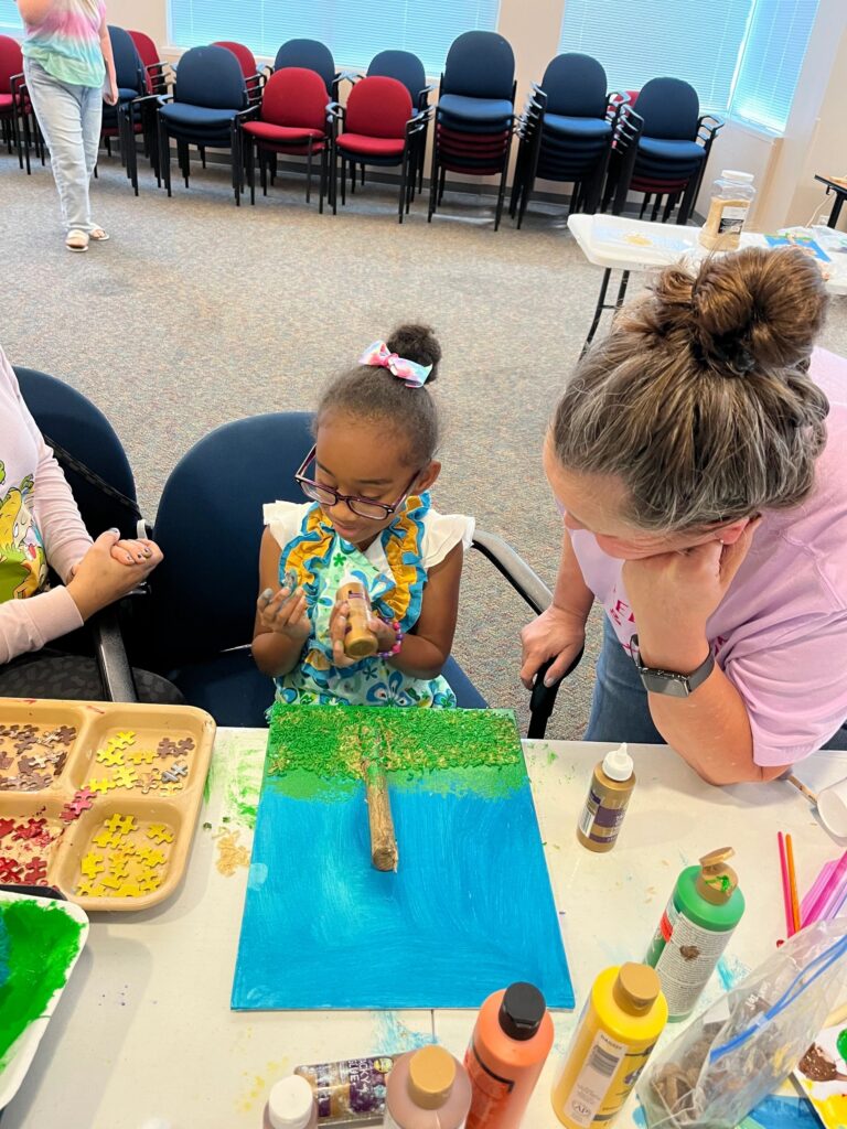 A volunteer helps an artist decorate a green and blue canvas with a twig laid across it, at a table with paint bottles and a divided tray nearby.