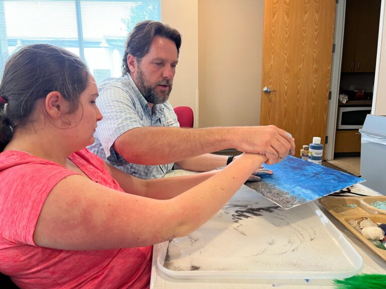 A volunteer helps an artist press their hands into a sand-textured area on a blue canvas, with shells and blue gravel in a tray on the table.