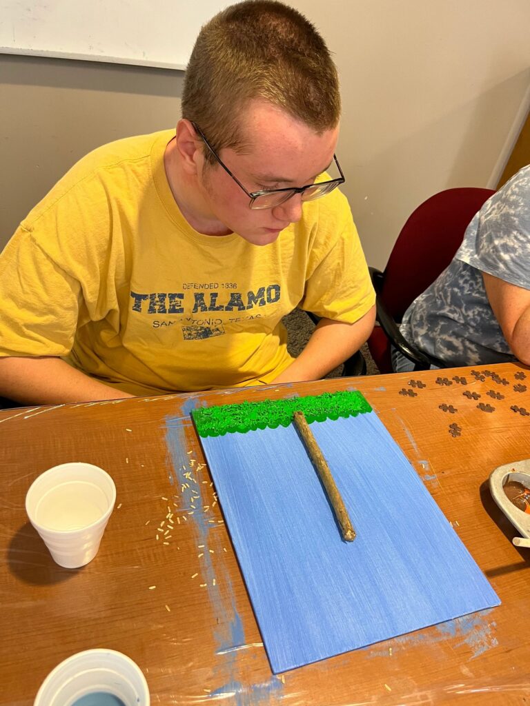 An artist paints a blue-green canvas with a strip of green sprinkled grass and a twig laid across the top, working at a table.