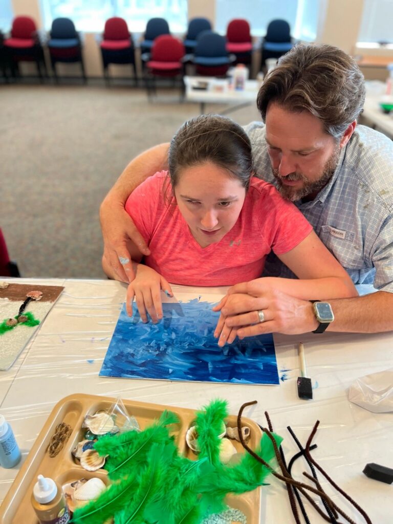 A volunteer helps an artist trace fingers across a finished dark blue canvas, with a tray of shells and green feathers on the table.