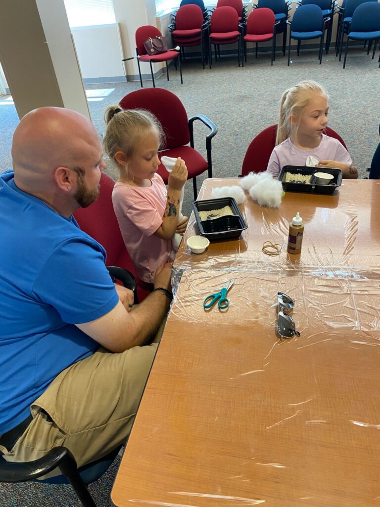 A volunteer looks on as a young artist holds a small blue jar of beads, with bowls of craft supplies and twine spools on the table.