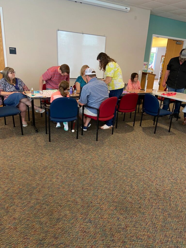A wide view of the studio, with artists and volunteers seated around several tables working on painted bottles and paper flowers.