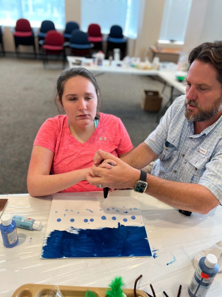 A volunteer helps an artist paint a row of light blue dots above a dark blue painted band on a canvas, with a green pom-pom charm nearby.