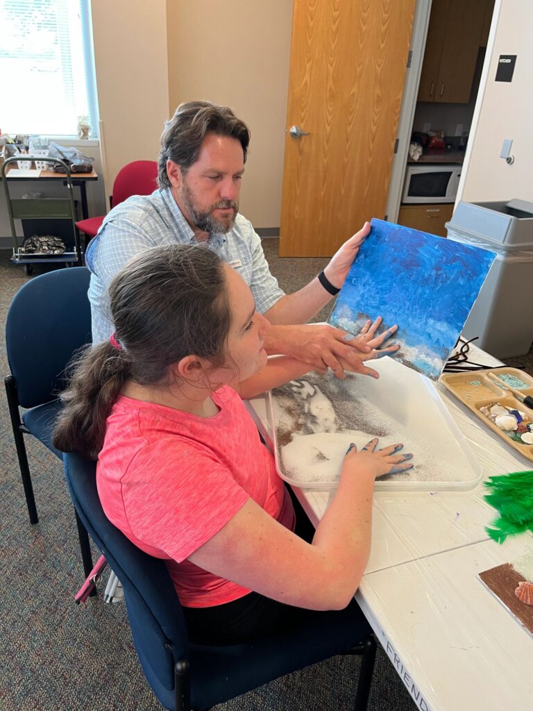 A volunteer helps an artist press their hands into sand on a blue canvas, with a tray of shells and green feathers on the table.