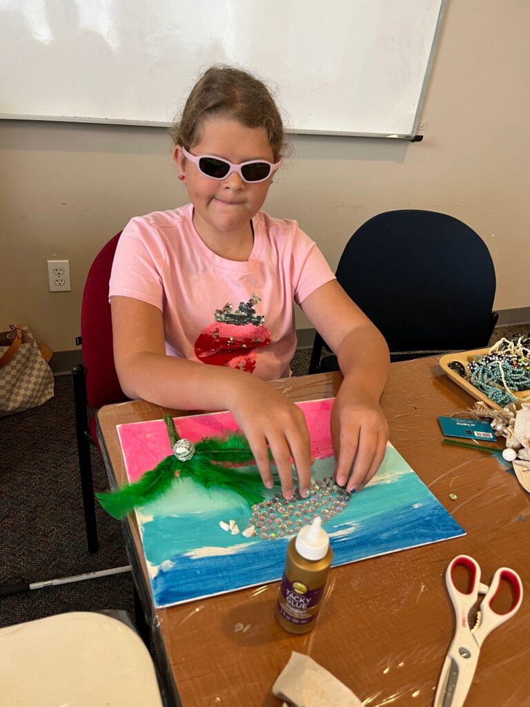 An artist glues green feathers and small rhinestones onto a pink and turquoise canvas, with tacky glue and scissors on the table.