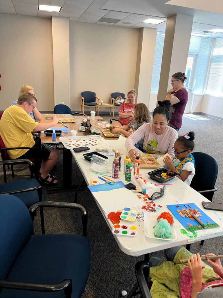 A wide view of the studio, with artists and volunteers working at a long table covered in paint trays, puzzle pieces and canvases in progress.