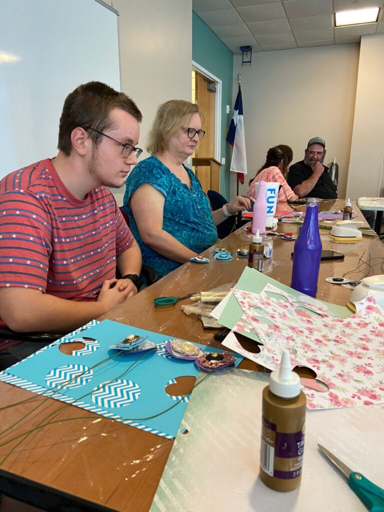 A young artist wearing sunglasses and a red bandanna sits with a boy at a table, a whiteboard and a support cane visible behind them.