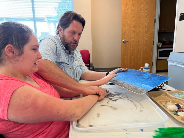 A volunteer helps an artist press their hands into a sand-textured area on a blue canvas, with a plate of shells and blue gravel on the table.