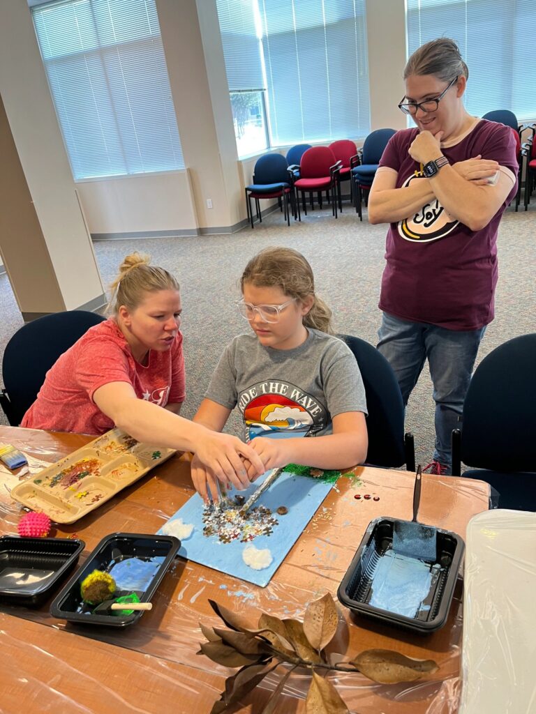 A volunteer helps an artist press puzzle pieces and small gems into a canvas painted with a twig, sky and grass, at a table with paint trays and dried leaves nearby.